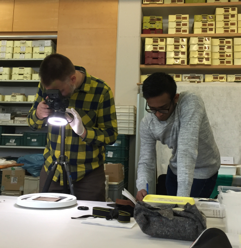 Professor wearing a yellow and black plaid shirt uses a ring light on a DSLR camera to image a wooden tablet sitting on the table. Undergraduate student in a grey sweatshirt and glasses is standing next to the professor taking notes next to him.