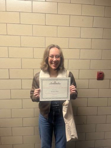 Student, Kirsten Bell stands in front of a white brick wall holding an award certificate