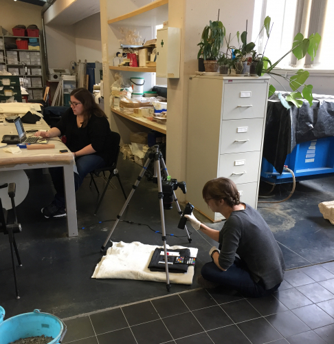 Student wearing black sweater and jeans is running the capture program for RTI on a laptop at a desk while a woman in a blue sweater and jeans sits on the floor with the RTI set-up and manages the flash.