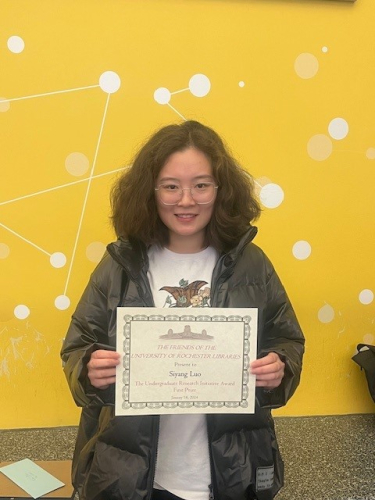 Student, Siyang Luo stands in front of a yellow wall holding an award certificate