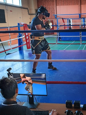 A person wearing a VR headset and boxing gloves trains in a boxing ring while someone monitors their progress on a screen nearby.